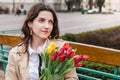 Beautiful young woman with spring tulips flowers bouquet at city street. Happy girl sitting on a bench outdoors. Spring portrait Royalty Free Stock Photo
