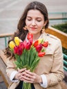 Beautiful young woman with spring tulips flowers bouquet at city street. Happy girl sitting on a bench outdoors. Spring portrait Royalty Free Stock Photo