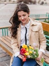 Beautiful young woman with spring tulips flowers bouquet at city street. Happy girl sitting on a bench outdoors. Spring portrait Royalty Free Stock Photo