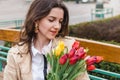 Beautiful young woman with spring tulips flowers bouquet at city street. Happy girl sitting on a bench outdoors. Spring portrait Royalty Free Stock Photo