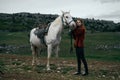 Beautiful young woman standing in the mountains and holding a white horse, nature