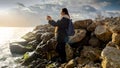 Beautiful young woman standing on the rocks at sea coast and making photographs of cold sea waves using smartphone. Concept of Royalty Free Stock Photo