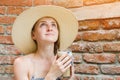 Beautiful young woman in straw hat holding cocktail and looking thoughtfully upwards. Cute girl on brick wall background Royalty Free Stock Photo