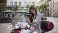 Beautiful young woman in stylish glasses holding vintage camera and sitting on the modern scooter in the middle of the