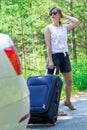 Beautiful young woman with a suitcase by the car on a forest road on a summer sunny day against the backdrop of green trees Royalty Free Stock Photo