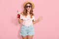 Beautiful young woman in summer blouse, short, hat and sunglasses, holding jug with cold beverage while standing on pink