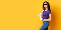 Beautiful young woman in sunglasses, purple shirt, blue jeans posing with bag on the wonderful yellow background