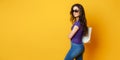 Beautiful young woman in sunglasses, purple shirt, blue jeans posing with bag on the wonderful yellow background