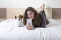 Beautiful young woman taking a selfie with mobile phone on bed with her cute small dog besides. Home, indoors and lifestyle Royalty Free Stock Photo