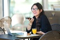 Beautiful young woman talking with her mobile phone while having breakfast while working with her laptop in a coffee shop. Royalty Free Stock Photo