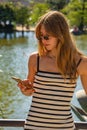 Beautiful young woman in a tight white dress with black stripes and long hair typing a message with her mobile phone with a lake Royalty Free Stock Photo