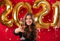 Beautiful young woman toasting with glass of champagne on New Year party over red studio background with gold balloons Royalty Free Stock Photo