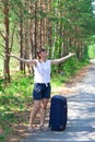 Beautiful young woman tourist with a suitcase on a forest road on a summer sunny day against the backdrop of green trees Royalty Free Stock Photo