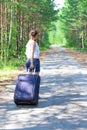 Beautiful young woman tourist with a suitcase on a forest road on a summer sunny day against the backdrop of green trees Royalty Free Stock Photo