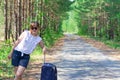 Beautiful young woman tourist with a suitcase on a forest road on a summer sunny day against the backdrop of green trees Royalty Free Stock Photo