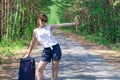 Beautiful young woman tourist with a suitcase on a forest road on a summer sunny day against the backdrop of green trees Royalty Free Stock Photo