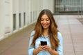 Beautiful young woman is using an app in her smartphone device to send a text message while standing in courtyard of office blocks