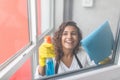 Beautiful young woman is using a duster and a spray, looking at camera and smiling while cleaning windows in the house Royalty Free Stock Photo