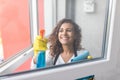Beautiful young woman is using a duster and a spray, looking at camera and smiling while cleaning windows in the house Royalty Free Stock Photo