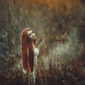 A beautiful young woman with very long red hair as a witch walks through the autumn forest.