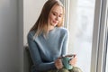 Beautiful young woman in warm sweater drinking hot tea on windowsill at home Royalty Free Stock Photo