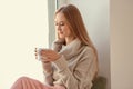 Beautiful young woman in warm sweater drinking hot tea on windowsill at home Royalty Free Stock Photo