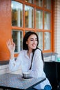 A beautiful young woman waves to passers-by at a table in a cafe. A young pretty girl wants to make an order