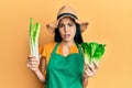 Beautiful young woman wearing gardener apron holding vegetables clueless and confused expression