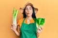 Beautiful young woman wearing gardener apron holding vegetables making fish face with mouth and squinting eyes, crazy and comical