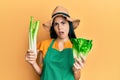 Beautiful young woman wearing gardener apron holding vegetables in shock face, looking skeptical and sarcastic, surprised with
