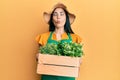 Beautiful young woman wearing gardener apron holding wooden plant pot making fish face with mouth and squinting eyes, crazy and