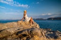 Beautiful young woman wearing traw hat sitting in rocks in white dress by the seashore