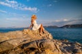 Beautiful young woman wearing traw hat sitting in rocks in white dress by the seashore