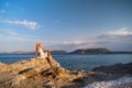 Beautiful young woman wearing traw hat sitting in rocks in white dress by the seashore