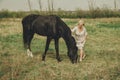 A beautiful young woman in a white chiffon dress with a short blonde haircut strokes and plays with a black horse in a meadow Royalty Free Stock Photo