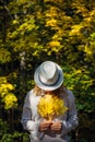 Beautiful young woman in white clothes and hat tilted her head to the bouquet of yellow leaves in her hands. Elegant blonde, face