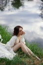 Beautiful young woman in white dress resting on shore lake pond river. Professional makeup and hairstyle with curled hair Royalty Free Stock Photo