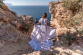 A beautiful young woman in a white light dress with long legs stands on the edge of a cliff above the sea waving a white Royalty Free Stock Photo