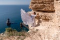 A beautiful young woman in a white light dress with long legs stands on the edge of a cliff above the sea waving a white Royalty Free Stock Photo