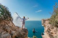 A beautiful young woman in a white light dress with long legs stands on the edge of a cliff above the sea waving a white Royalty Free Stock Photo