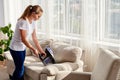 Beautiful young woman in white shirt and jeans cleaning sofa with vacuum cleaner in living room, copy space. Housework.