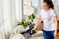 Beautiful young woman in white shirt and jeans cleaning sofa with vacuum cleaner in living room, copy space. Housework.