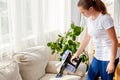 Beautiful young woman in white shirt and jeans cleaning sofa with vacuum cleaner in living room, copy space. Housework.