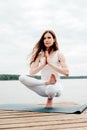 Beautiful young woman in white sports clothes is sitting on  wooden pier on lake in  balance yoga position with namaste gesture Royalty Free Stock Photo
