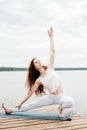 Beautiful young woman in white sports clothes in warrior yoga pose on wooden pierce on lake outdoor