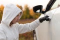 A beautiful young woman in a white sweater with a hood and black gloves opens the car door to steal it. Selective focus