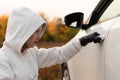 A beautiful young woman in a white sweater with a hood and black gloves opens the car door to steal it. Selective focus