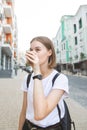 Beautiful young woman in a white T-shirt and a backpack drinks coffee from a glass on the street Royalty Free Stock Photo