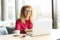 Beautiful young woman working remotely on her fashionable laptop at hipster coffee shop. Happy female freelancer with trendy wrist Royalty Free Stock Photo