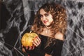 Beautifull woman in a witch`s costume is holding pumpkin in front of black wall with spider net